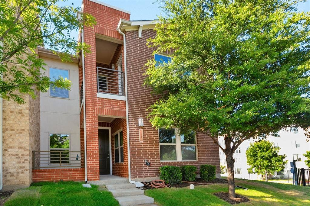 Contemporary facade featuring red brick and stucco, a prominent front entry, upper-level balcony with metal railing, and a ground-level patio with matching railing