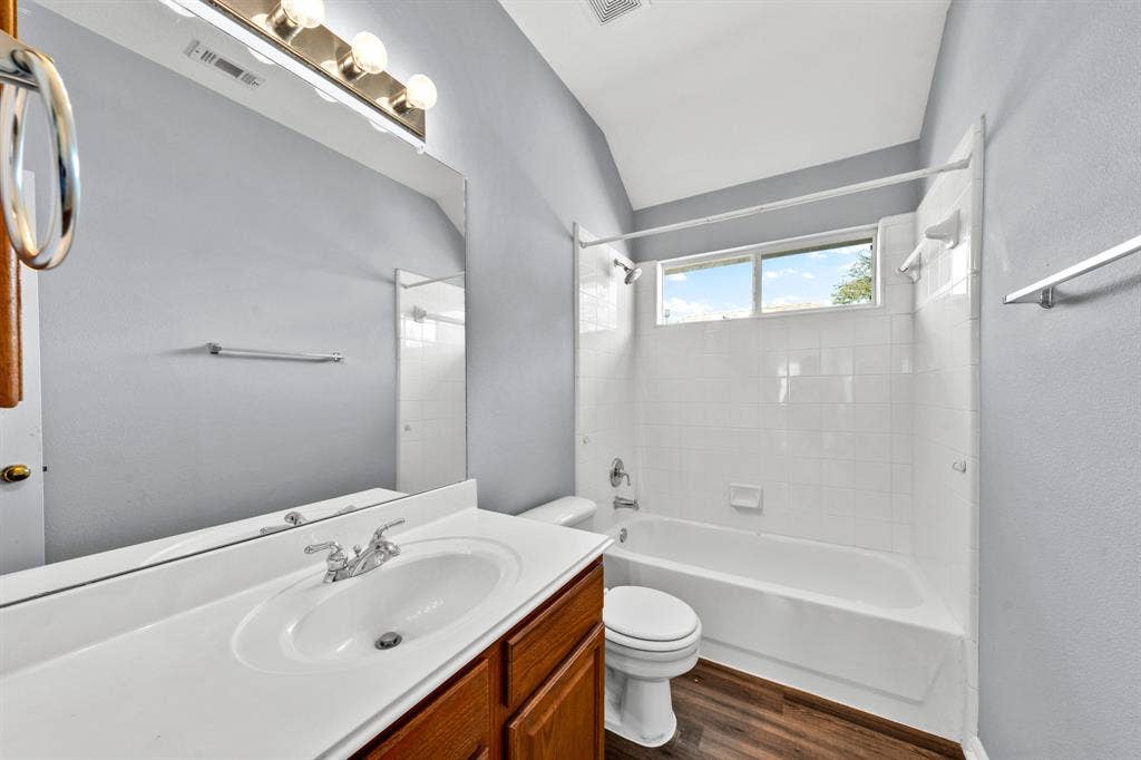 Bathroom featuring a single vanity with wood-finish cabinetry, a large frameless wall mirror, a bathtub with a shower, and wood-finish flooring