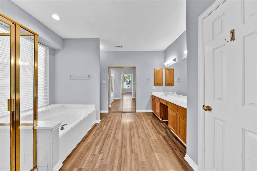 Integrated soaking tub with tiled surround, wood-finish flooring, dual vanity with wood cabinetry, full-height mirrored closet doors, and recessed lighting