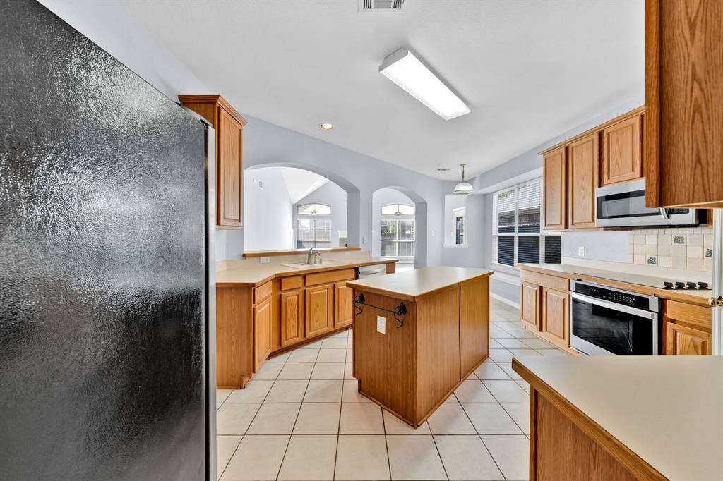 Kitchen featuring wood cabinetry, a center island, built-in stainless steel microwave and oven, a white tile backsplash, and tile flooring