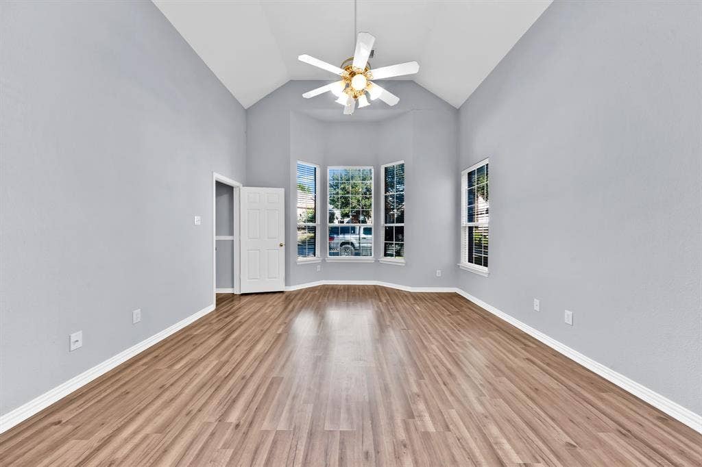 Vaulted ceiling, wood-finish flooring, bay window, white trim, and a ceiling fan with integrated lighting