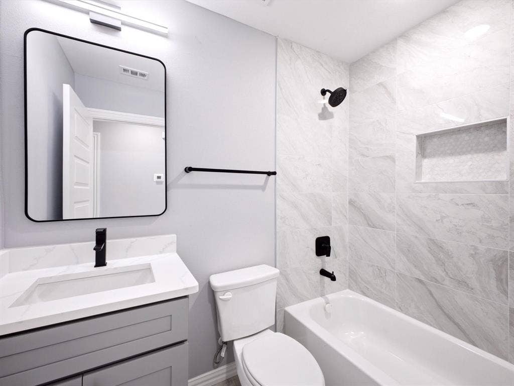 1727 Mentor Avenue, Dallas, TX 75216 White tiled tub and shower enclosure with a recessed shelf, black shower fixtures, a gray vanity with an integrated sink, and a black-framed mirror