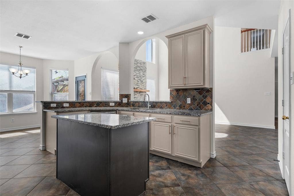 Expansive kitchen featuring a central island with a stone-finish countertop, coordinating perimeter countertops, light cabinetry, and a mosaic tile backsplash