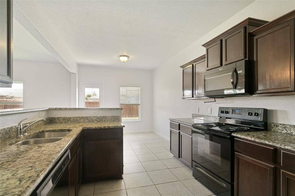 Kitchen with dark wood cabinetry, light speckled countertops, a double basin stainless steel sink, tile flooring, and integrated black appliances