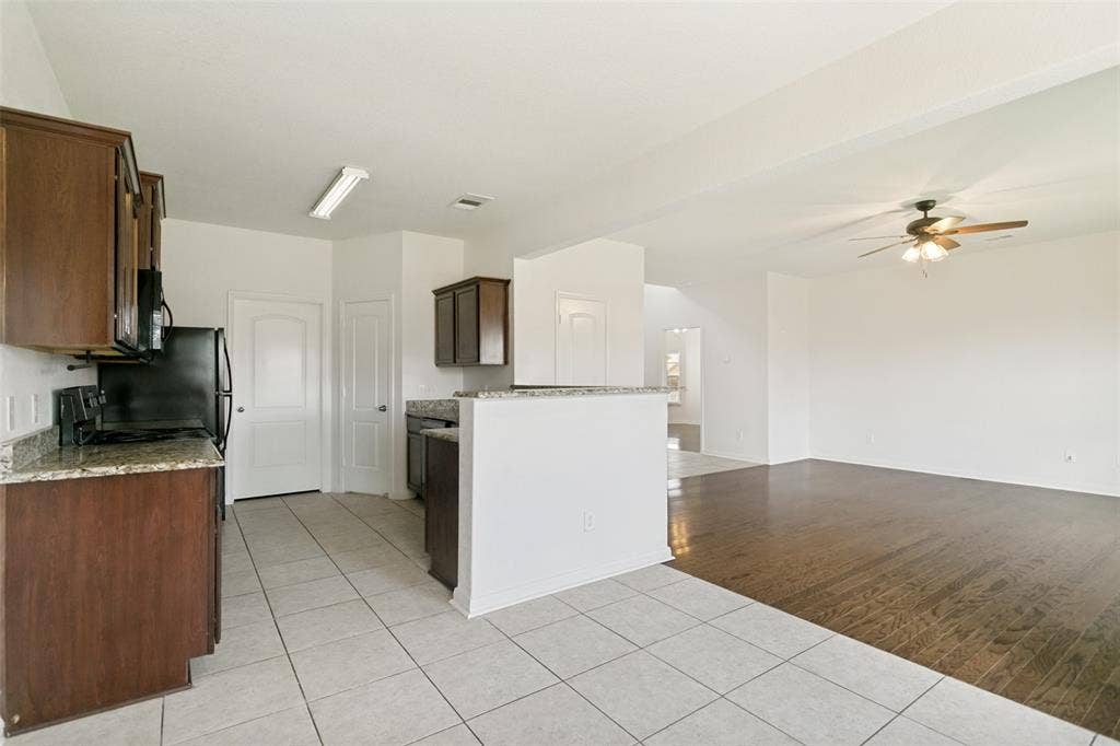 Kitchen featuring tile flooring, granite-style countertops, wood-finish cabinetry, a built-in microwave, and an open concept layout to the adjacent living area with wood-finish flooring and a ceiling fan
