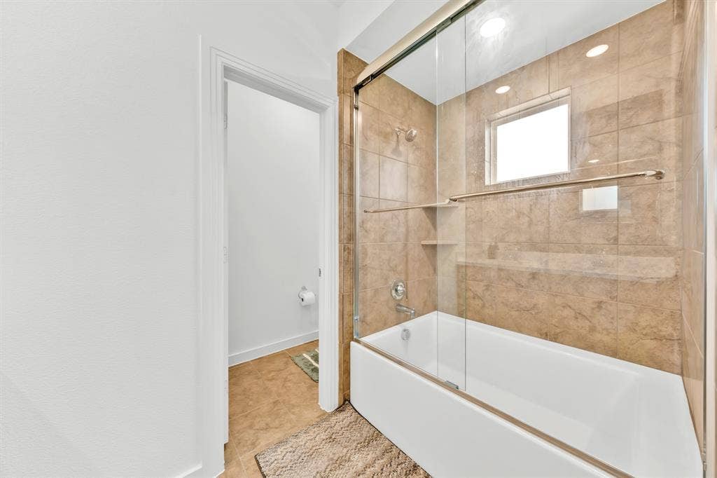 3928 Bison Lane, Crandall, TX 75114 White bathtub with a sliding glass enclosure, stone-finish tile surround, built-in shower niche, and a frosted window