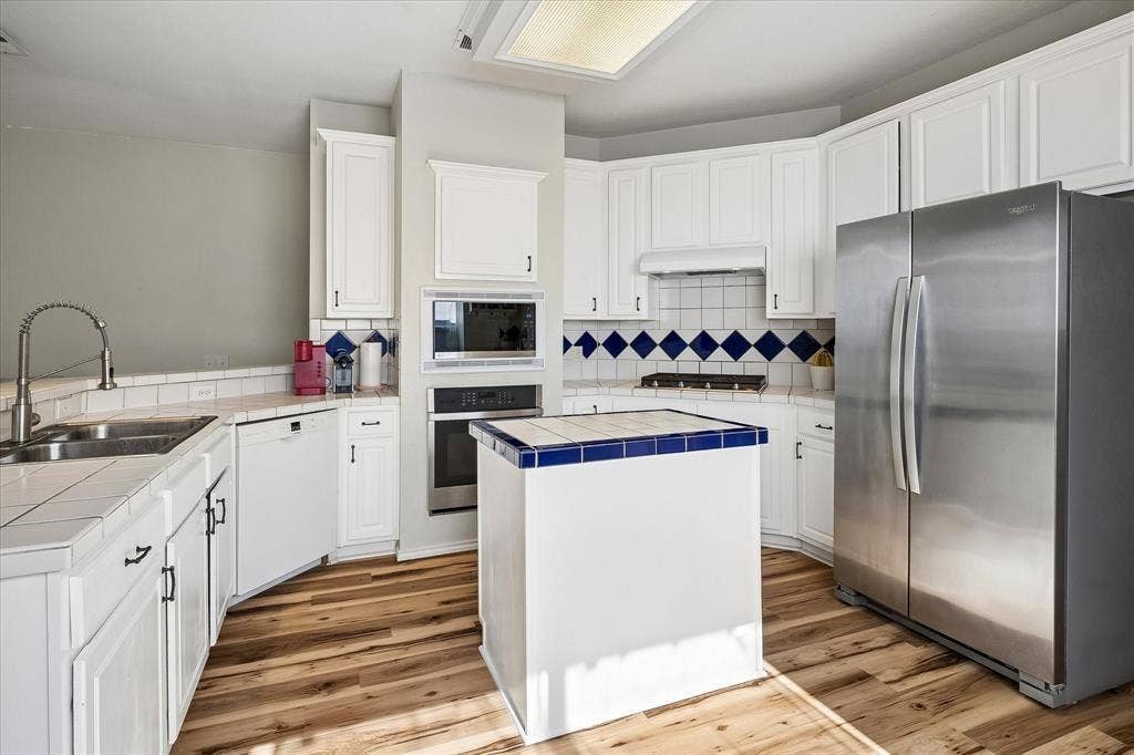 11379 Lockshire Drive, Frisco, TX 75035 Kitchen featuring white cabinetry, tile countertops, wood-finish flooring, built-in stainless steel appliances, and a central island with tile trim