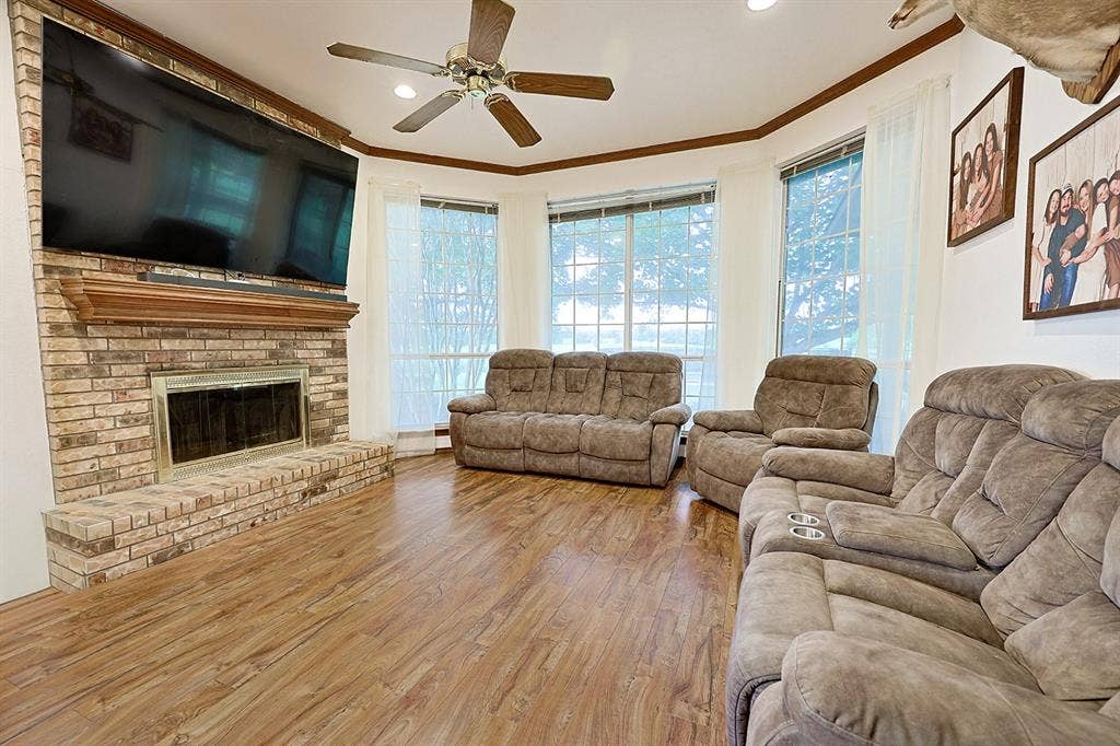 2042 County Road 1072, Greenville, TX 75401 Brick fireplace surround with a wood mantel, wood-finish flooring, bay window configuration, and a ceiling fan with integrated lighting
