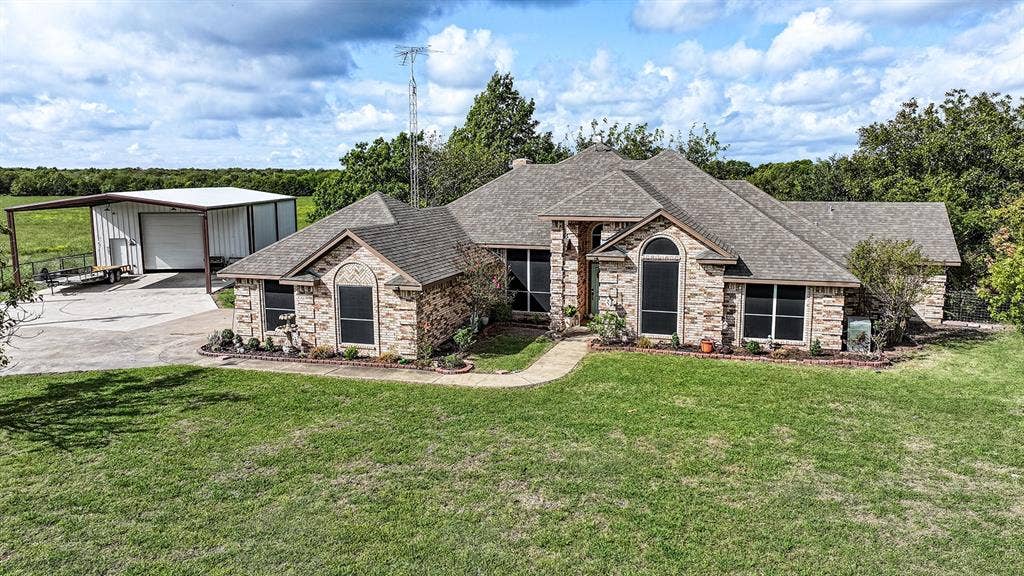 2042 County Road 1072, Greenville, TX 75401 Single-story masonry exterior featuring a multi-gabled roofline, concrete driveway, attached metal-framed workshop, and landscaping beds