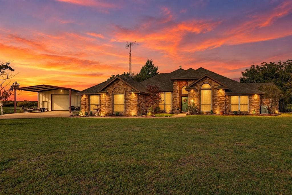2042 County Road 1072, Greenville, TX 75401 Stone masonry exterior featuring an arched entryway, shingle roof, expansive front lawn, and a metal-roofed detached structure