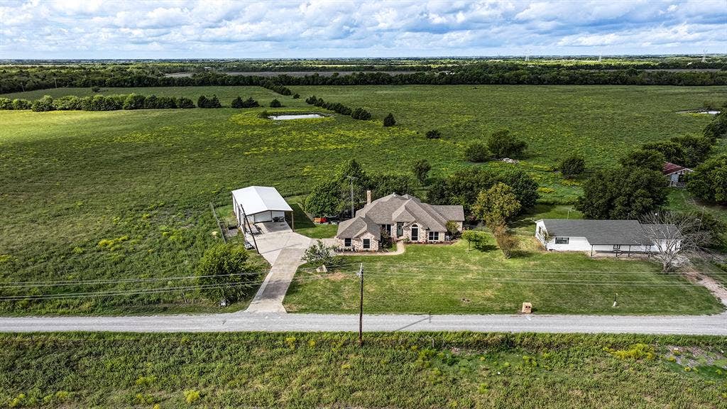 2042 County Road 1072, Greenville, TX 75401 Aerial perspective of a multi-structure property featuring a main residence with a pitched roof, a detached metal-sided workshop, and a secondary structure with a covered porch, all situated on expansive acreage
