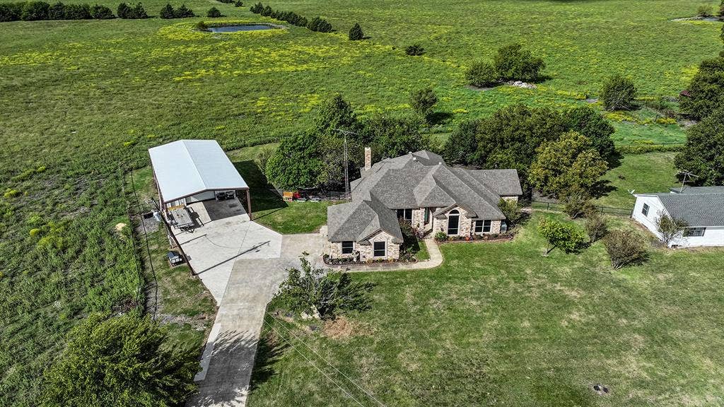 2042 County Road 1072, Greenville, TX 75401 Aerial view of a sprawling property featuring a multi-gabled residence with stone masonry, an expansive concrete driveway, a detached metal-roofed carport, and fenced pasture grounds