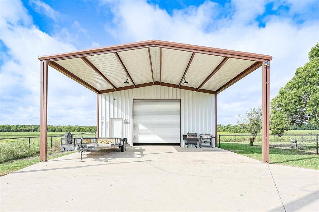 2042 County Road 1072, Greenville, TX 75401 Metal-sided detached structure featuring a high-clearance carport, concrete foundation, roll-up overhead door, and integrated lighting