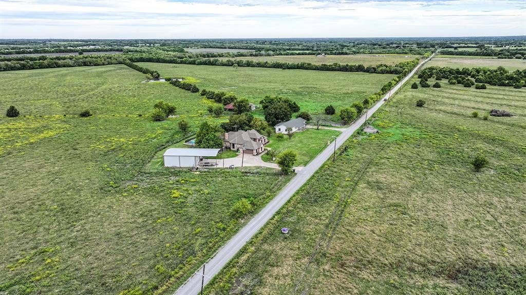 2042 County Road 1072, Greenville, TX 75401 Aerial perspective of a multi-structure property featuring expansive acreage, a residential dwelling, a detached metal-sided workshop, and a paved driveway