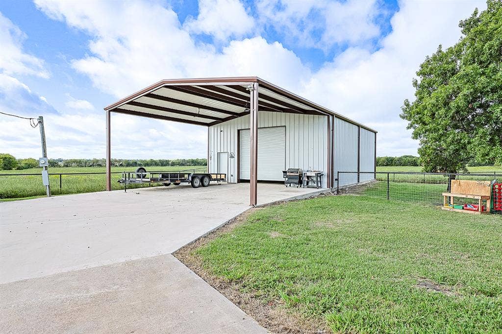 2042 County Road 1072, Greenville, TX 75401 Metal-sided workshop building with a concrete parking pad, oversized roll-up door, side entry door, and extended metal-roofed carport structure