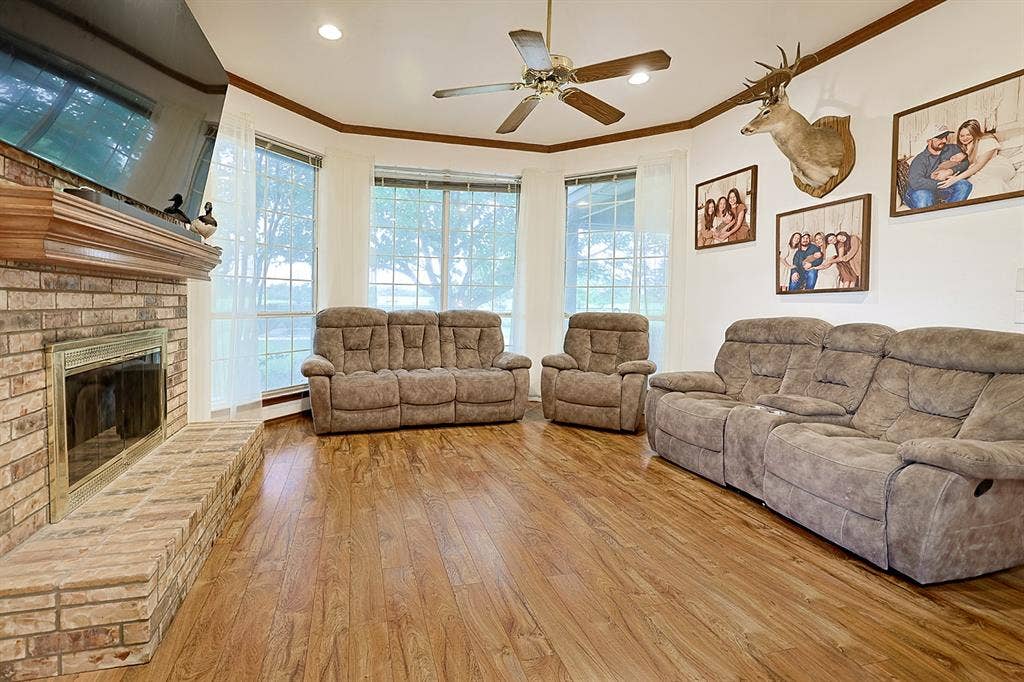 2042 County Road 1072, Greenville, TX 75401 Brick fireplace featuring a raised hearth and wooden mantel, wood-finish flooring, tray ceiling with a center fan, and multiple large windows