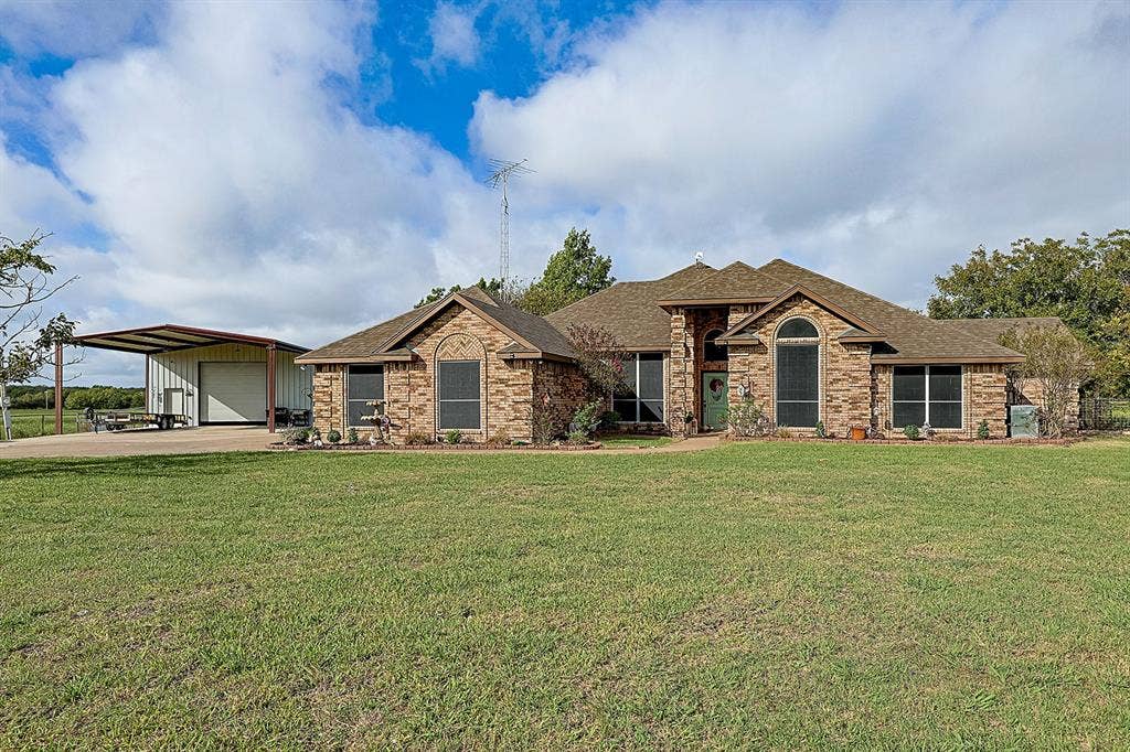 2042 County Road 1072, Greenville, TX 75401 Brick exterior residence featuring arched windows, a multi-gabled roofline, an expansive lawn, and a detached metal-frame workshop with a covered carport