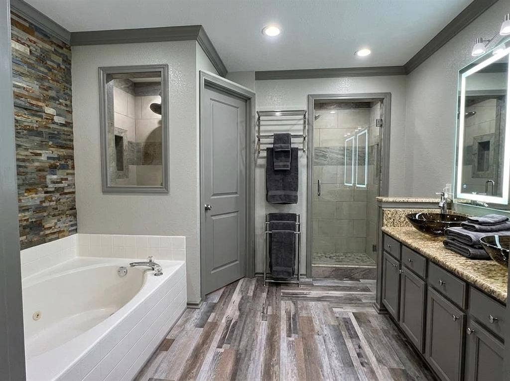 2500 Sierra Drive, McKinney, TX 75071 Soaking tub with tile surround, walk-in shower with glass enclosure, dual vanity with vessel sinks, wood-finish flooring, and a stacked stone accent wall