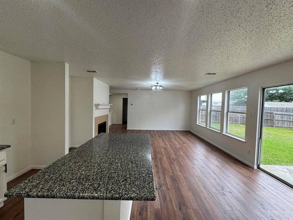 7305 Marsland Lane, Arlington, TX 76001 Kitchen island with a granite-style countertop, wood-finish flooring, a fireplace with mantel, multiple windows, and a sliding glass door opening to a fenced yard