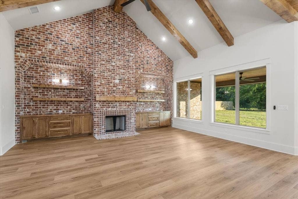 Vaulted ceiling with exposed wood beams, full-height brick accent wall with fireplace, built-in wooden cabinetry, wood-finish flooring, and large windows