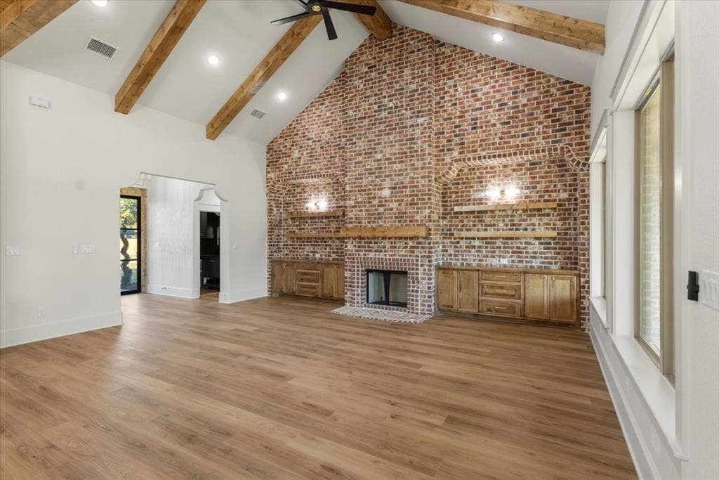 Vaulted ceilings with exposed wood beams, brick accent wall with fireplace, wood-finish flooring, built-in cabinetry, and large windows