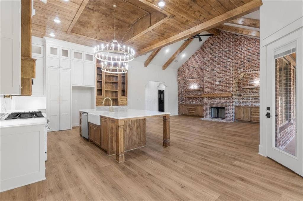 Vaulted wood-beamed ceiling with exposed trusses, wood-finish flooring, brick accent wall with fireplace, white shaker cabinetry, and a kitchen island with a farmhouse sink