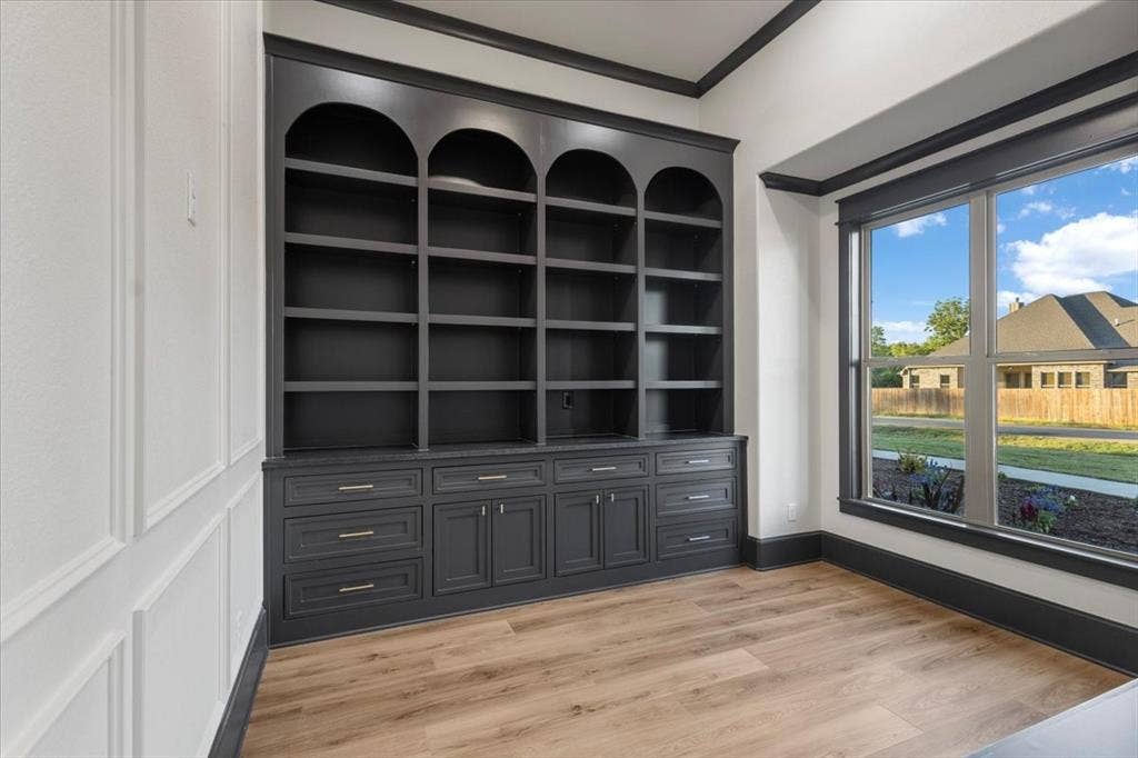 Built-in shelving with arched tops and lower cabinetry, wood-finish flooring, large window with dark trim, wainscoting detail on walls, and dark crown molding