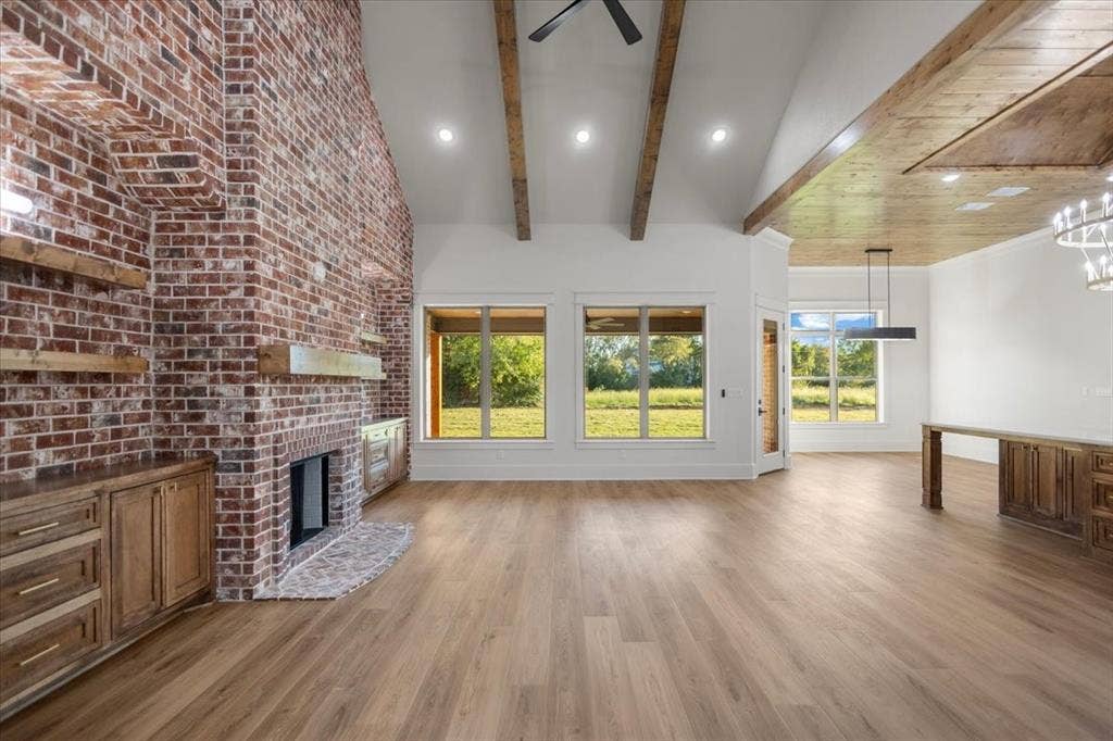 Soaring vaulted ceiling with exposed wood beams, brick fireplace with wood mantle, wood-finish flooring, recessed lighting, and large windows