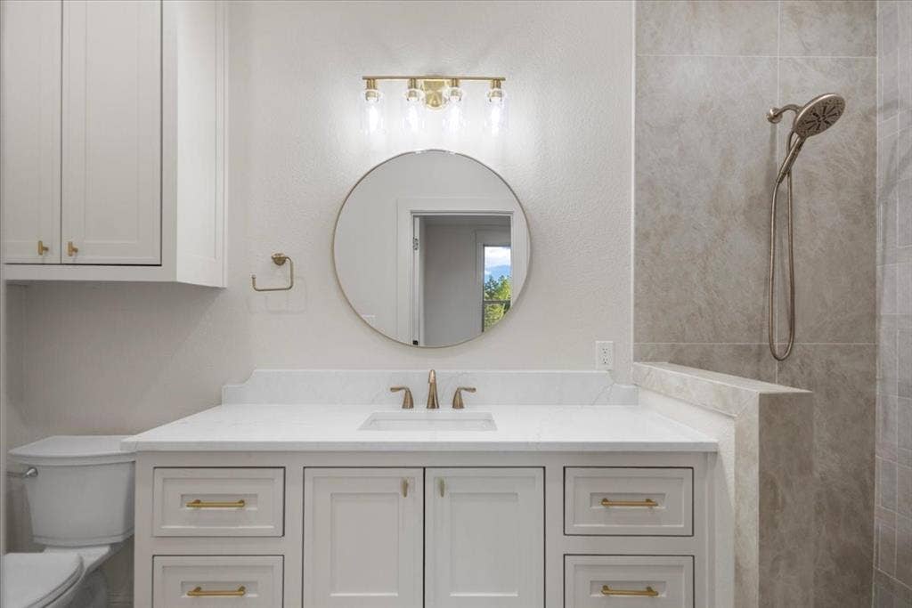 White shaker cabinetry with gold-finish hardware, a single vanity with an undermount sink, a round mirror, and a gold-finish vanity light fixture