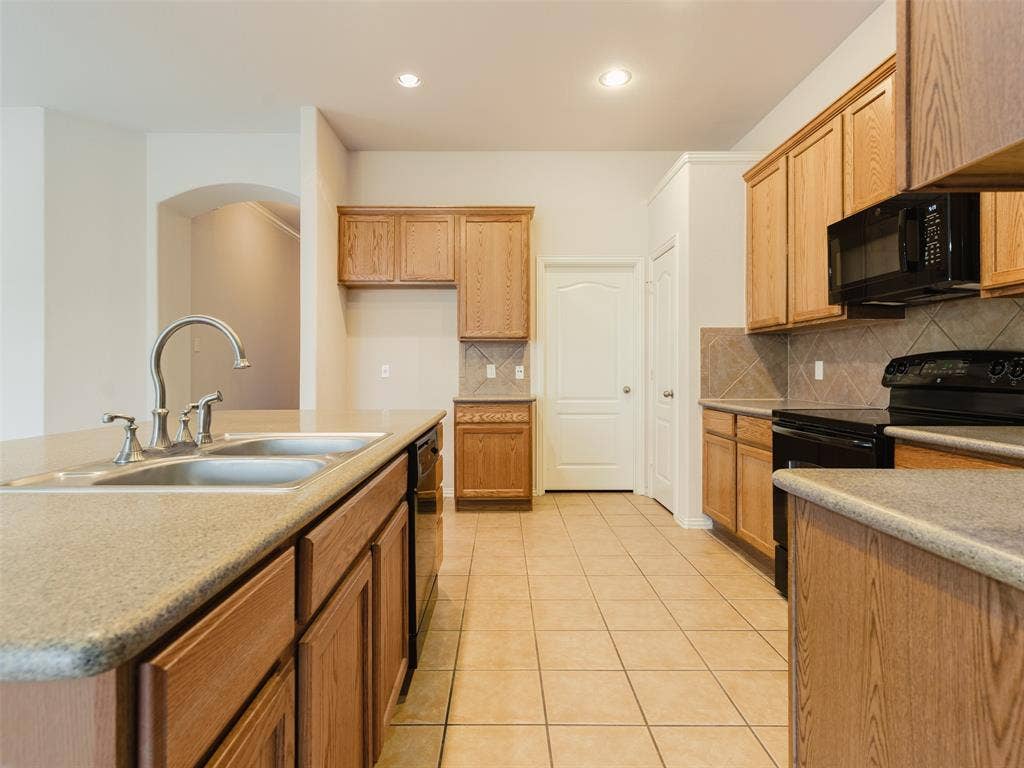 Kitchen featuring light wood cabinetry, stone-composite countertops, tile flooring, built-in black appliances, and a dual-basin stainless steel sink