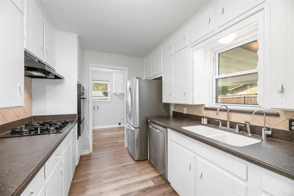 Galley kitchen featuring abundant white cabinetry, integrated double wall ovens, a gas cooktop with an overhead range hood, wood-finish flooring, and a double basin sink
