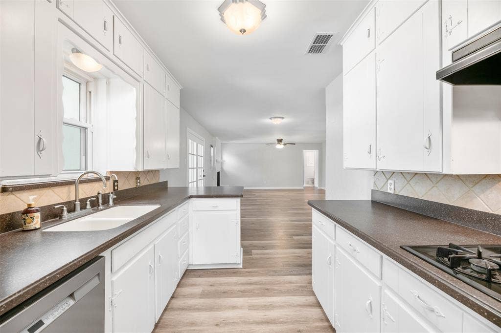Galley kitchen featuring white cabinetry, dark countertops, a double-basin sink, stainless steel dishwasher, and tile backsplash