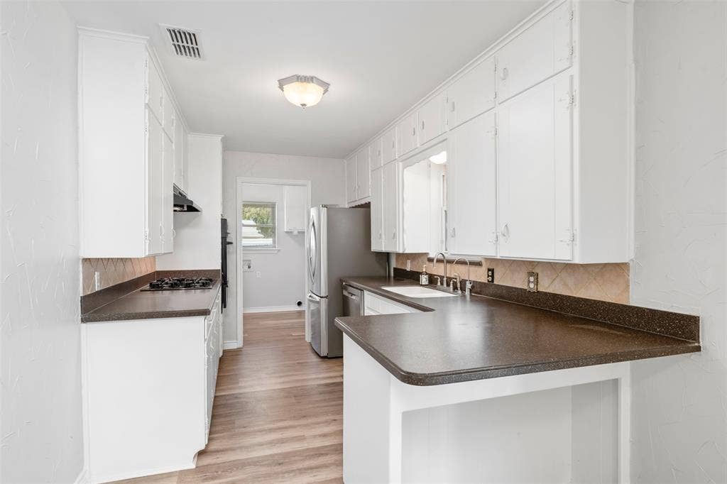 Galley kitchen featuring abundant white cabinetry, dark solid-surface countertops, a double basin stainless steel sink, and wood-finish flooring