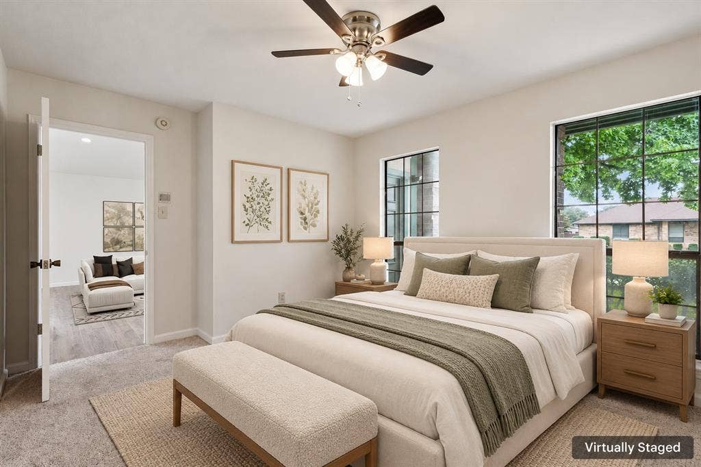 Carpeted bedroom with a ceiling fan, two windows featuring grid patterns, and a doorway opening to an adjacent room with wood-finish flooring