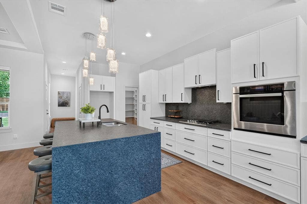 3210 Carpenter Avenue, Dallas, TX 75215 Kitchen featuring wood-finish flooring, white cabinetry with black hardware, dark stone countertops, a built-in wall oven, and a kitchen island with a sink and pendant lighting