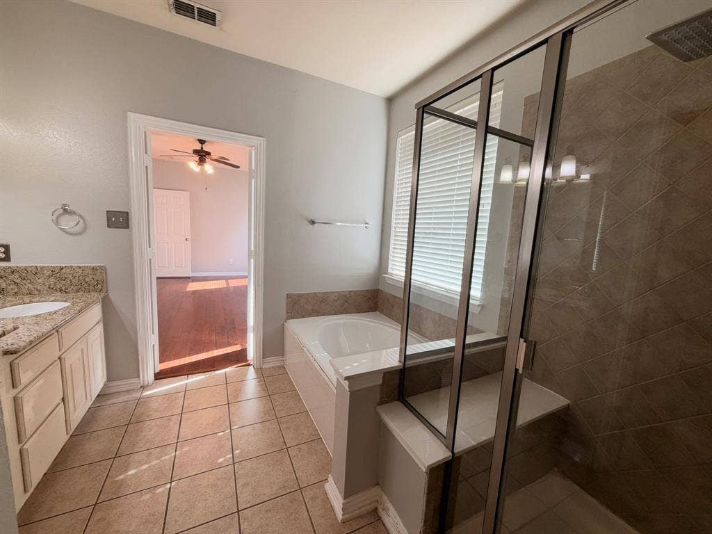 5016 Rushden Road, McKinney, TX 75070 Tiled flooring, dual vanity with stone countertops, built-in soaking tub, separate glass-enclosed shower with tiled surround, and a window with blinds