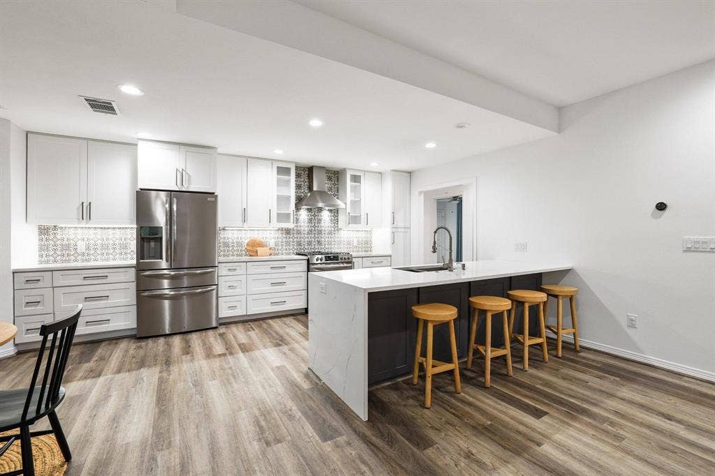 7668 La Cosa Drive, Dallas, TX 75248 Open-concept kitchen featuring white cabinetry, stainless steel appliances, patterned backsplash, an island with a built-in sink, and wood-finish flooring