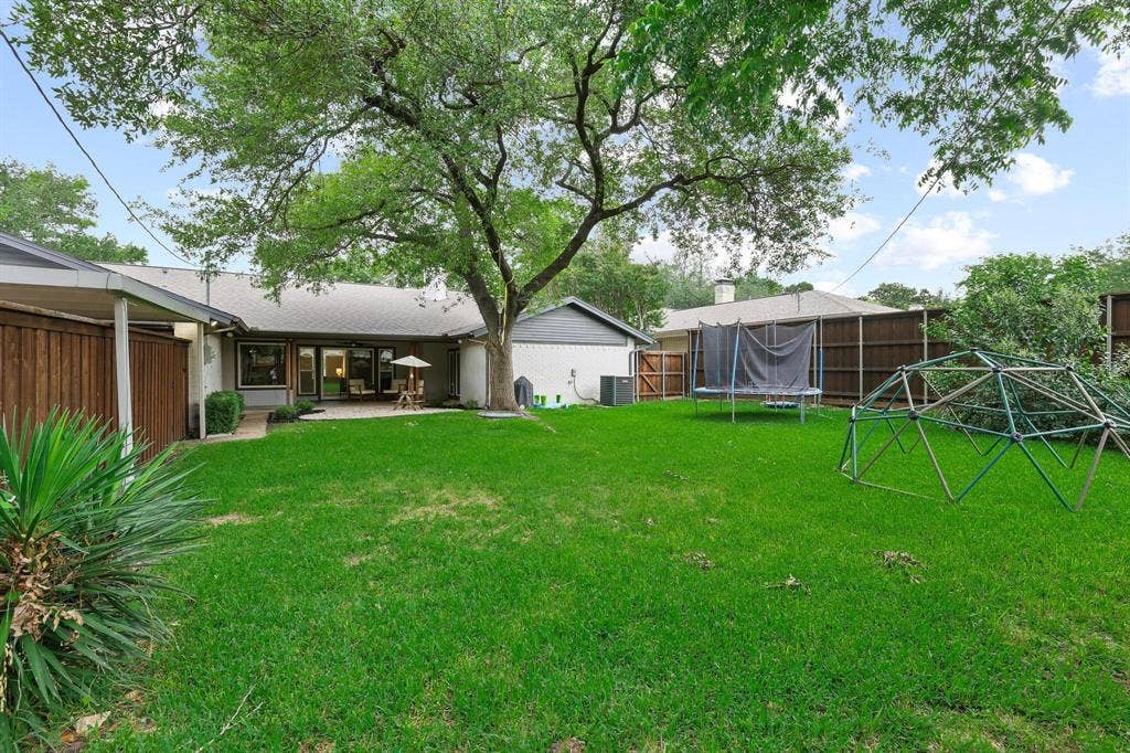 7668 La Cosa Drive, Dallas, TX 75248 Expansive backyard featuring a manicured lawn, mature shade tree, privacy fencing, and an outdoor patio