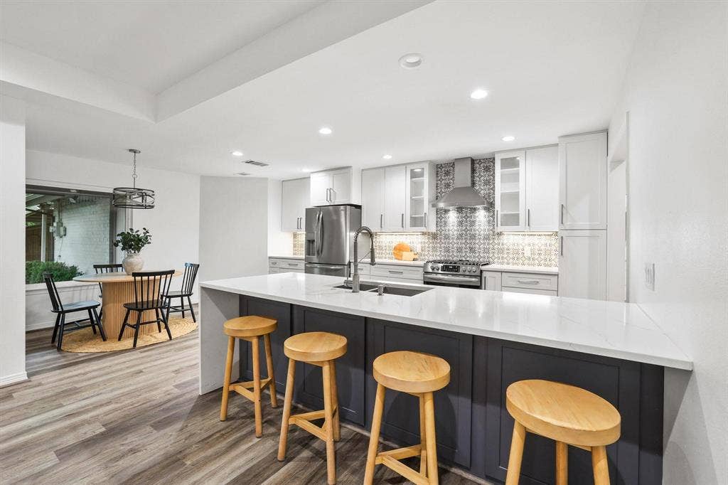 7668 La Cosa Drive, Dallas, TX 75248 Modern kitchen featuring a large island with a light-toned countertop, white cabinetry with glass inserts, patterned backsplash, and wood-finish flooring