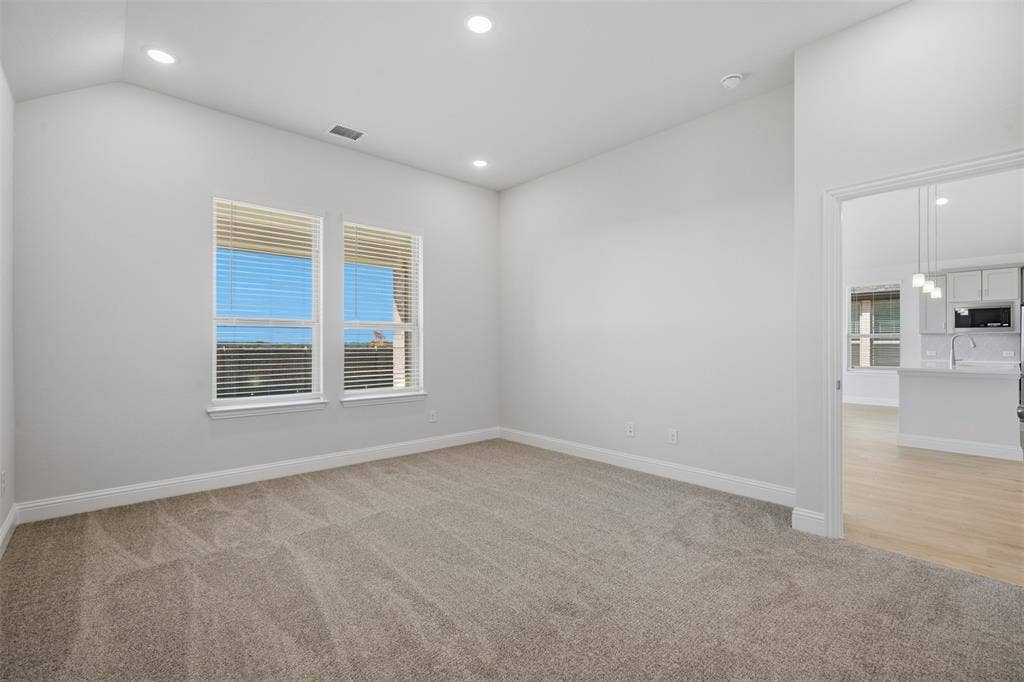 Vaulted ceiling with recessed lighting, two windows with blinds, plush carpet flooring, and a doorway to a kitchen with wood-finish flooring