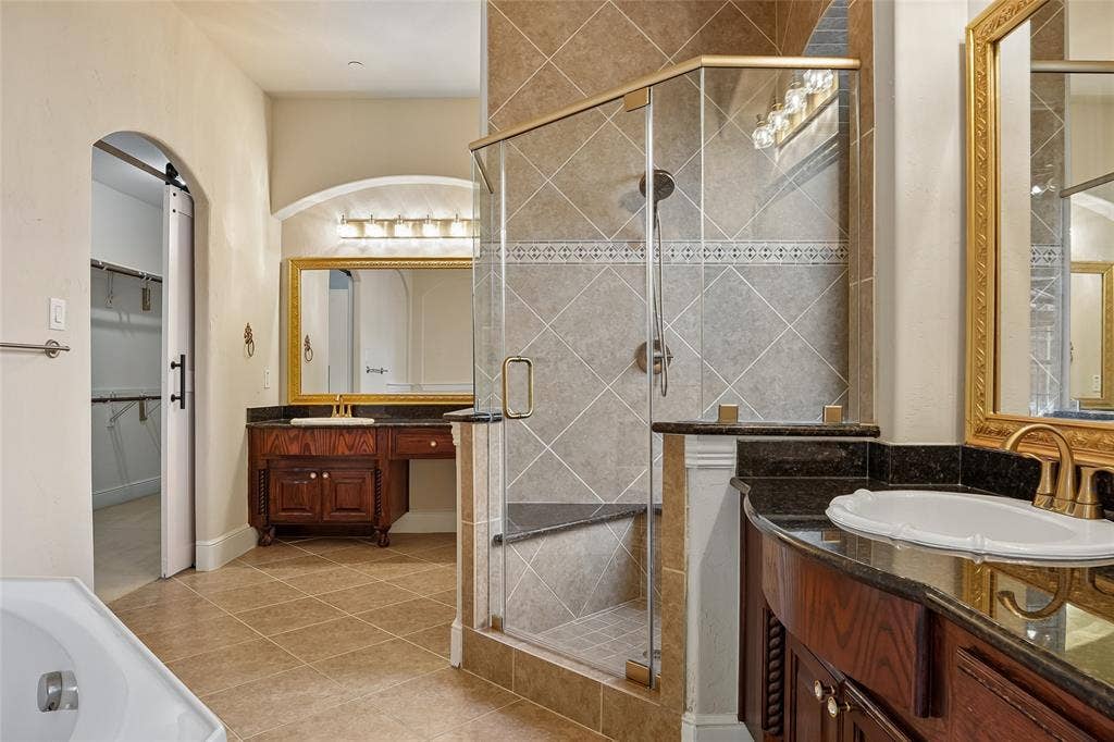 Walk-in shower with glass enclosure and built-in bench, dual vanity with undermount sinks and dark countertops, wood-finish cabinetry, and neutral tile flooring