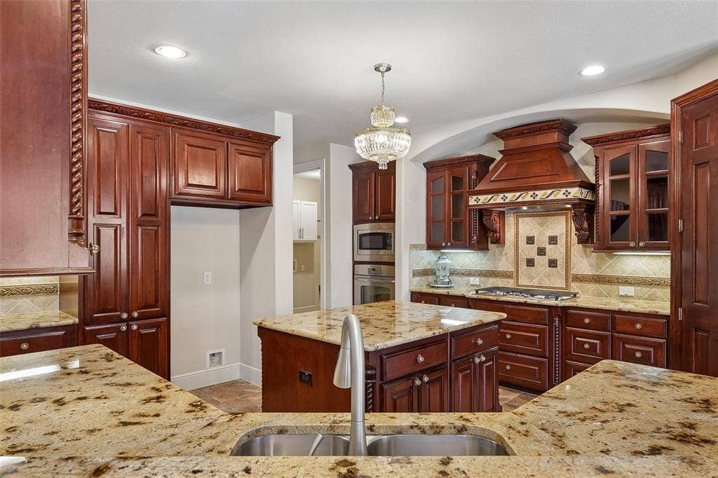 Spacious kitchen featuring rich wood cabinetry, granite countertops, a central island, stainless steel sink with a gooseneck faucet, and integrated stainless steel appliances