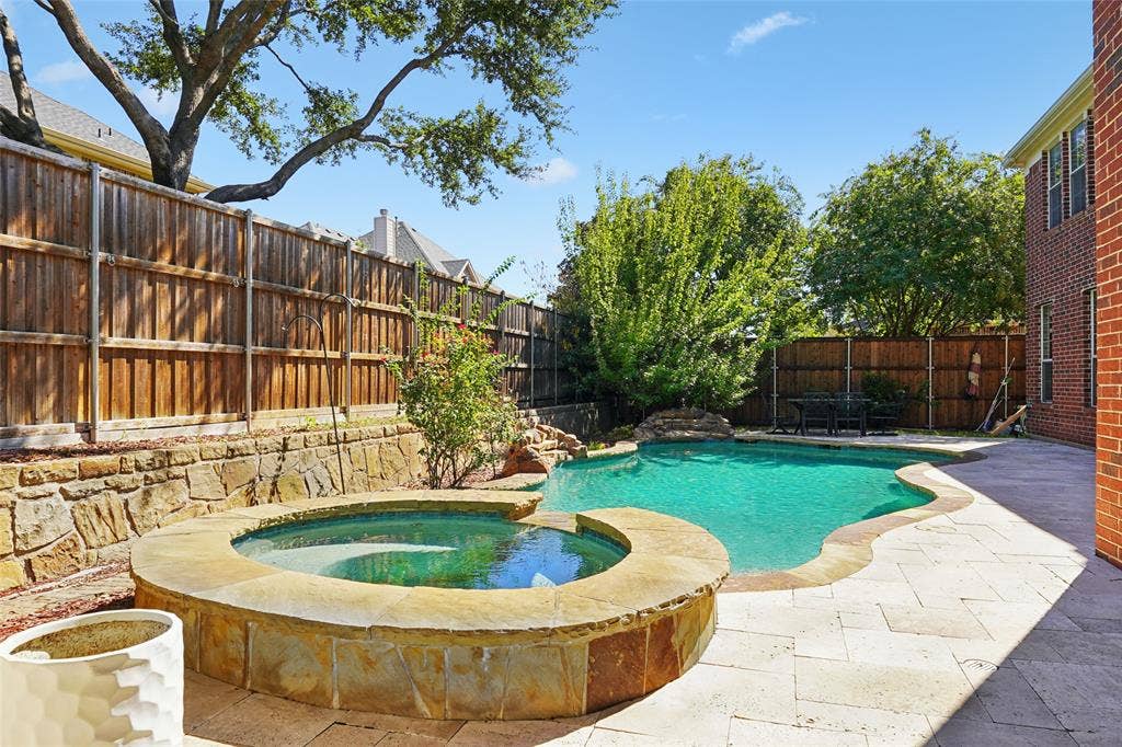 Backyard oasis featuring a curvilinear swimming pool, integrated spa with stone surround, natural stone coping, and a retaining wall with mature trees