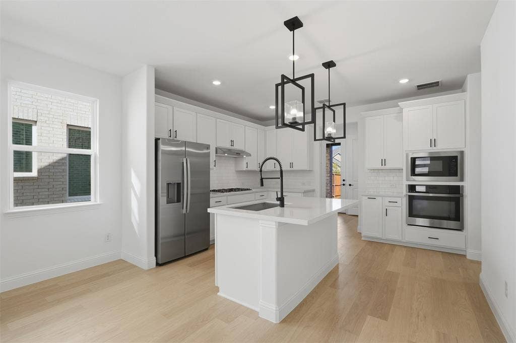 Open-concept kitchen featuring an island with a sink, stainless steel appliances, white cabinetry, wood-finish flooring, and two contemporary pendant lights