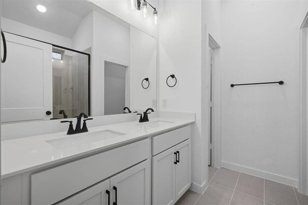 Dual white rectangular undermount sinks set in a light-toned countertop, white shaker-style cabinetry with matte black hardware, large wall-mounted mirror, and a clear glass shower enclosure with black framing