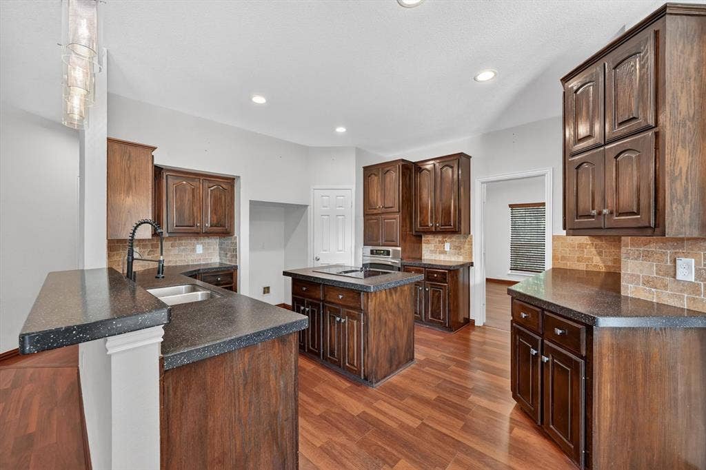512 Renfro Court, Irving, TX 75063 Kitchen featuring wood cabinetry, an island with an integrated cooktop, dark speckled countertops, brick-patterned tile backsplash, and wood-finish flooring