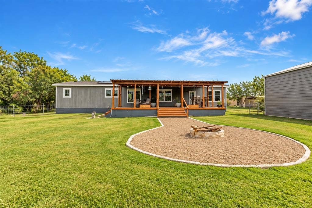 213 River Front Drive, Cedar Creek, TX 78612 Expansive backyard featuring a manicured lawn, a gravel fire pit area with stone surround, a large wood deck with pergola, and a detached building with metal siding