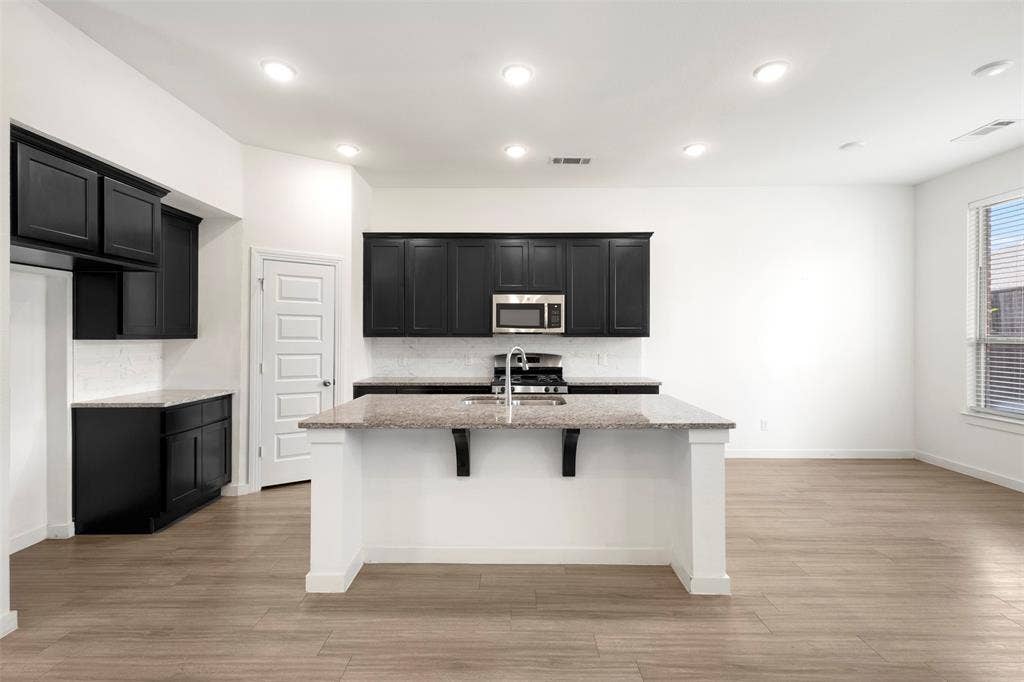 2466 San Marcos Drive, Forney, TX 75126 Kitchen featuring black shaker-style cabinetry, stone-surface center island with a stainless steel sink, wood-finish flooring, and recessed ceiling lighting