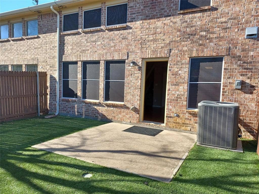 Brick exterior with multiple windows, a concrete patio, synthetic turf, and a wood privacy fence