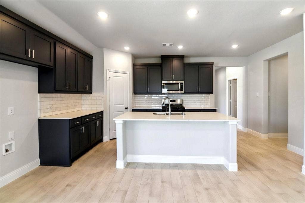 Kitchen featuring dark cabinetry, white subway tile backsplash, light countertops, a central island with a sink, and wood-finish flooring